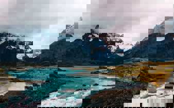 Turquoise glacial waters reflect rugged, snow-capped peaks beneath a moody sky in Torres del Paine National Park, Chile. 