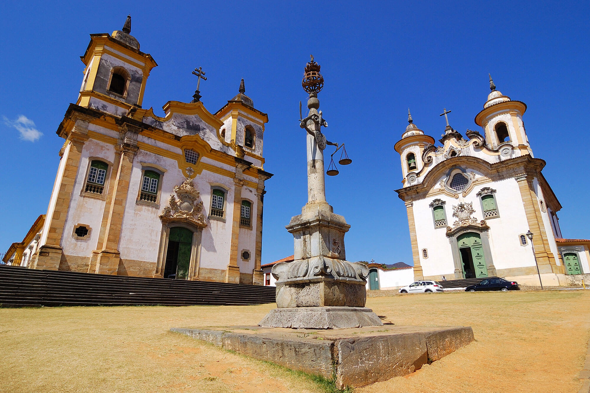 Twin colonial churches and monument in Tiradentes, a historic town in Minas Gerais, Brazil, under a clear blue sky. 