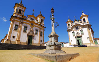 Twin colonial churches and monument in Tiradentes, a historic town in Minas Gerais, Brazil, under a clear blue sky. 