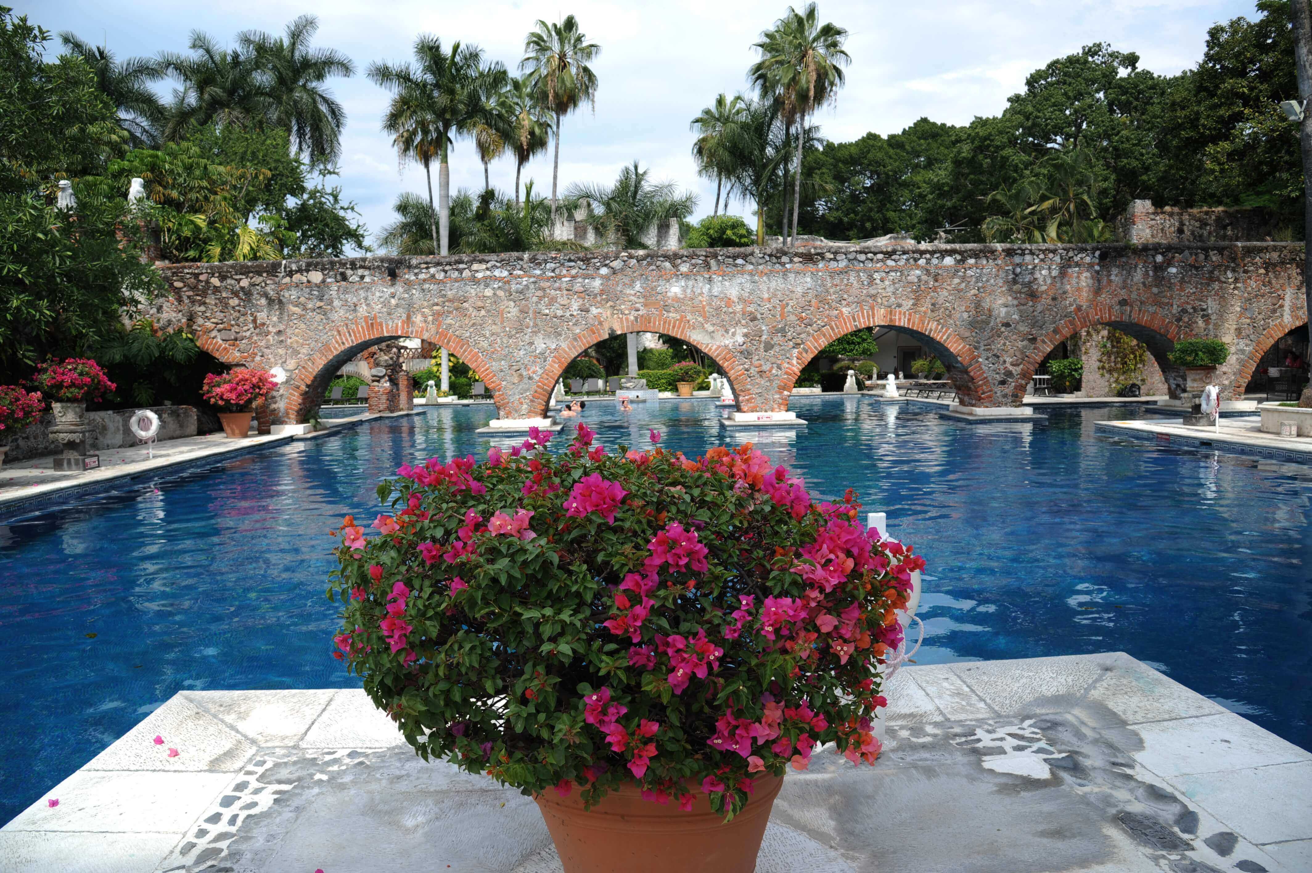 Charming Mexican hacienda with a serene pool, stone arches, and vibrant flowers in a tranquil garden setting. 